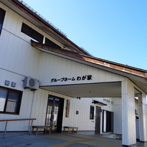 グループホームわが家の玄関・車寄せ。白い壁に看板があり、駐車スペース完備。